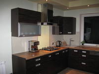 Ciemna, narożna zabudowa kuchenna z drewnianym blatem, widoczny ekspres do kawy i płyta gazowa, oświetlenie punktowe podszafkowe, okno w tle.